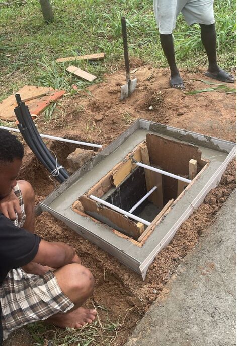 Overhead view of workers concreting a chamber collar with timber formwork, black conduit ducts led in from a trench and a shovel beside the pit