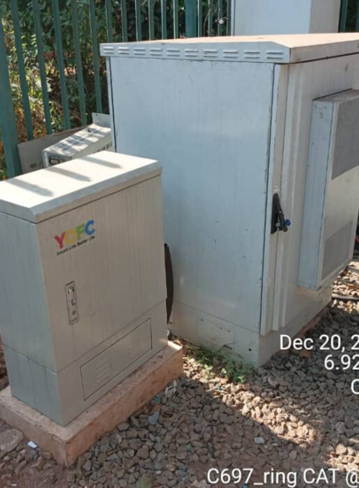 Two outdoor telecom cabinets set on concrete plinths over gravel in front of a green security fence